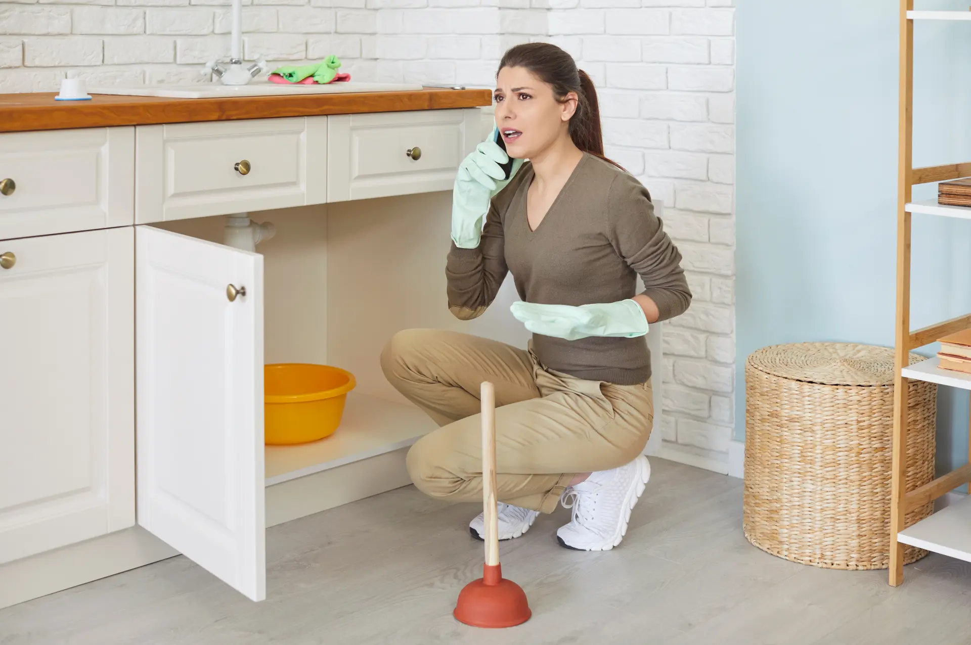 Žena rieši haváriu vody pod kuchynským drezom a telefonuje inštalatérovi pri úniku vody.